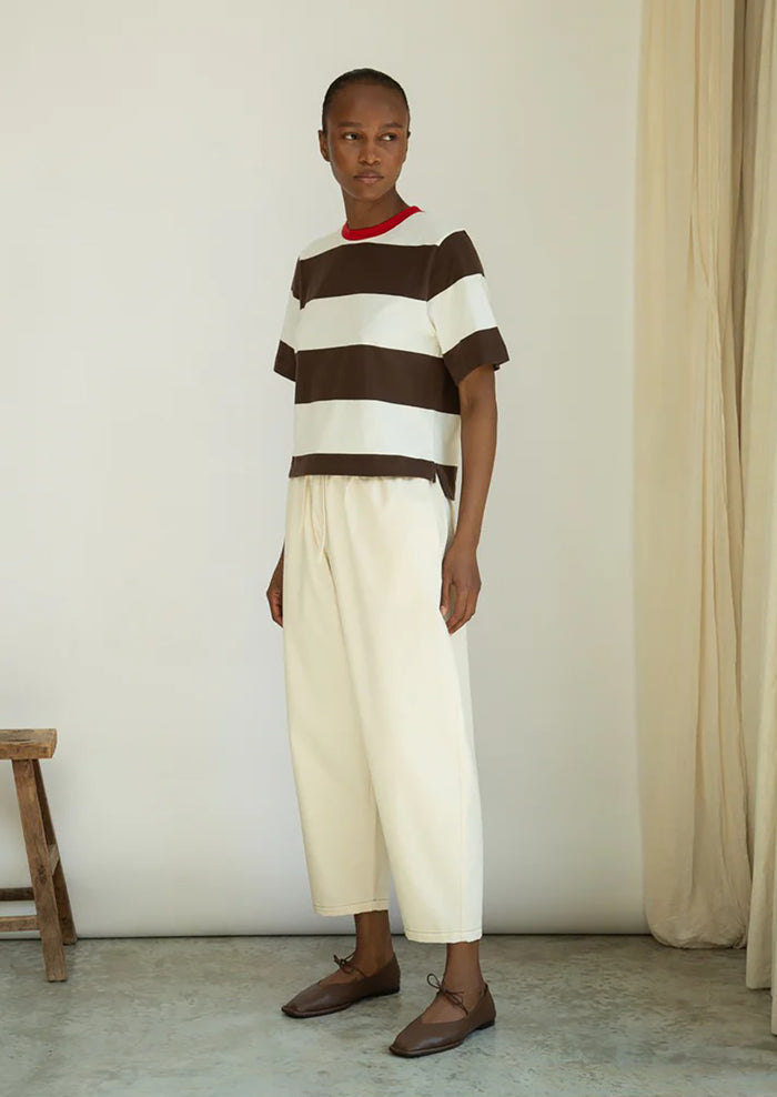 Person wearing a brown and white striped shirt with cream pants in a minimalistic setting.