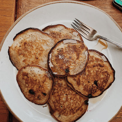 Six golden-brown pancakes with toasted edges arranged on a white ceramic plate with a beige rim, topped with a drizzle of syrup. A silver fork rests on the upper right side of the plate, which sits on a wooden table.