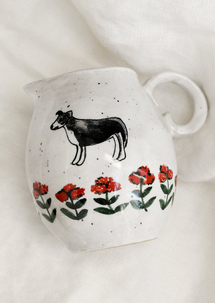 A small speckled white ceramic pitcher with illustration of dog with red flowers.