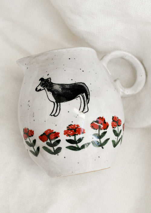 A small speckled white ceramic pitcher with illustration of dog with red flowers.