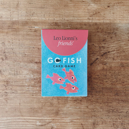Leo Lionni's Go Fish Card Game box, front view, teal and coral design with illustrated pink fish, flat lay on rustic wooden surface