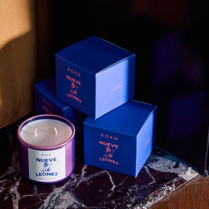 A Roen candle with a label 'NUEVE LEONES' displayed on a marble surface with blue product boxes stacked next to it.